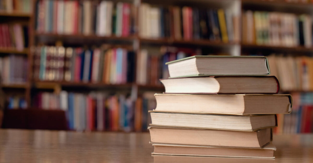 A pile of books on a table with filled bookshelves behind out of focus