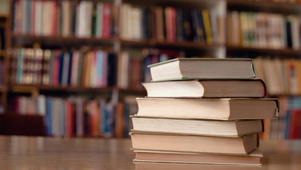 A pile of books on a table with filled bookshelves behind out of focus