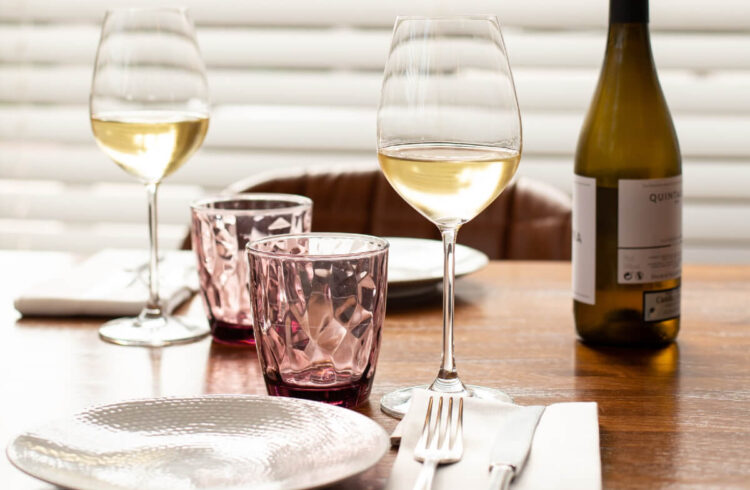 A bottle and two glasses of white wine on a dinner table with water glasses, plate and cutlery