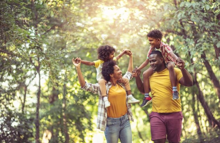 A smiling family walk through a sunny woodland the children on their parents shoulders