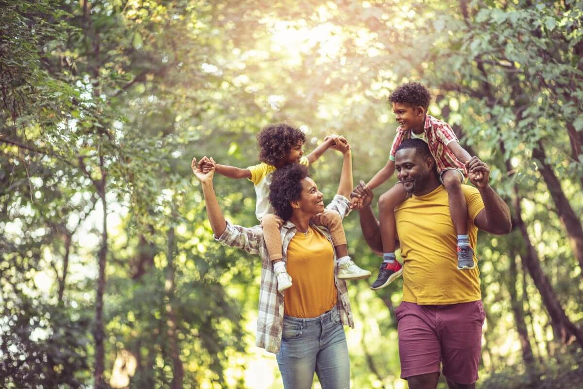 A smiling family walk through a sunny woodland the children on their parents shoulders