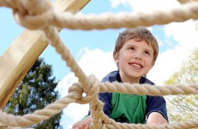 A child smiles and climbs on a rope frame at Witley Court Play Area