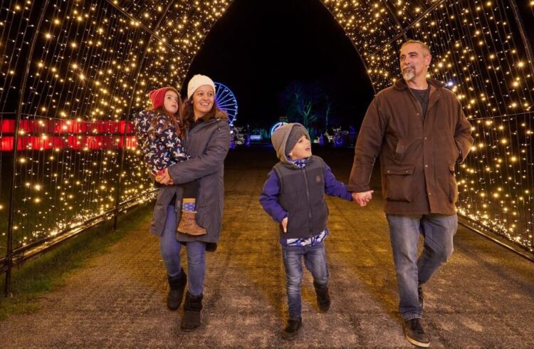 A family group (two adults and two children) walk through the light trail at Winter Glow