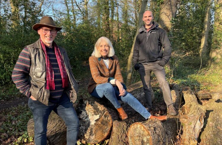 Three people pose in a wooded area upon a pile of logs