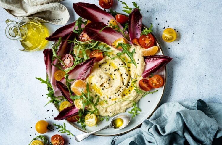 a plate of hummus decorated with purple lettuce leaves and tomatoes on a white marble surface with linen napkins.