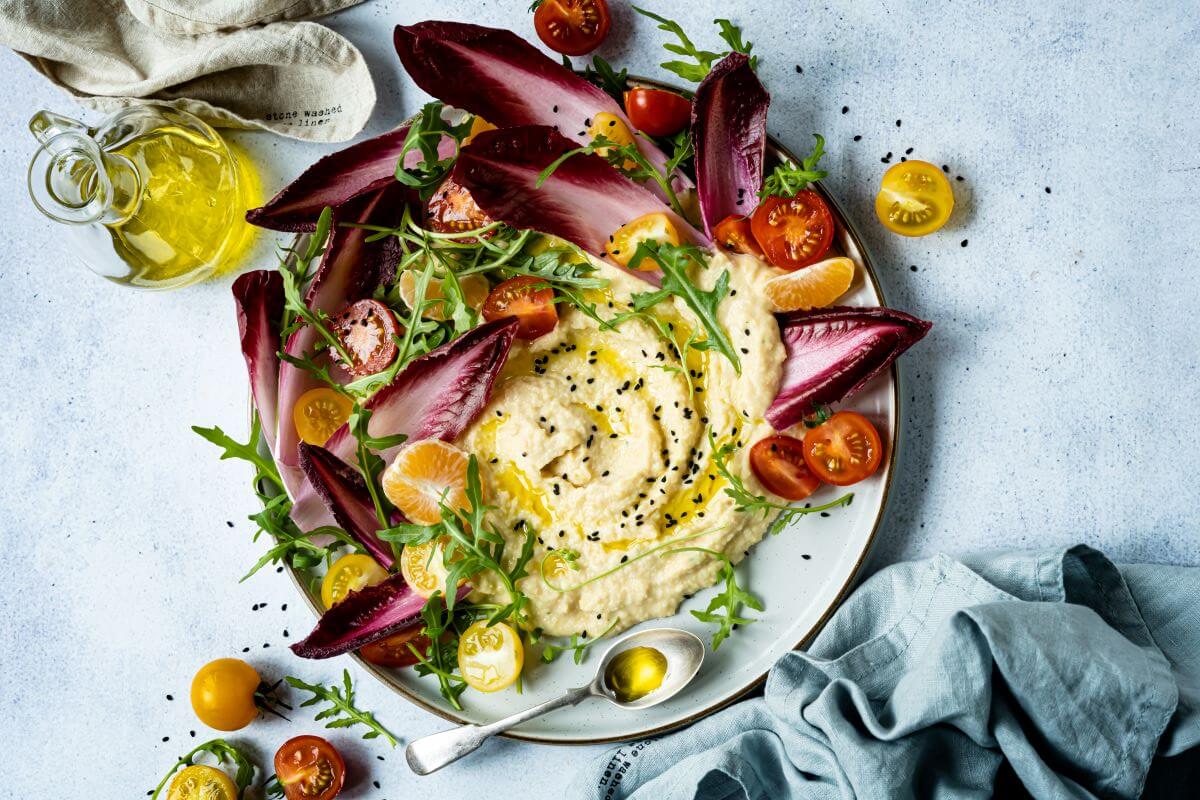 a plate of hummus decorated with purple lettuce leaves and tomatoes on a white marble surface with linen napkins.