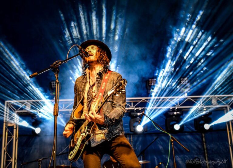 A man plays electric guitar and sings onstage with stage lights in the background