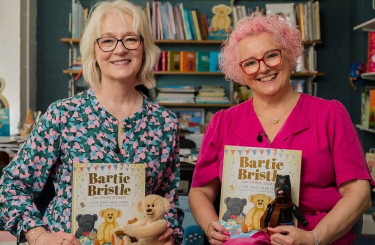 Two authors hold their book and a teddy bear. They are smiling warmly.