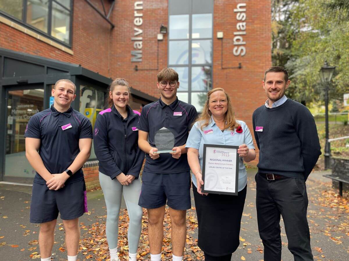 five people stand outside a brick leisure centre holding a certificate and smiling